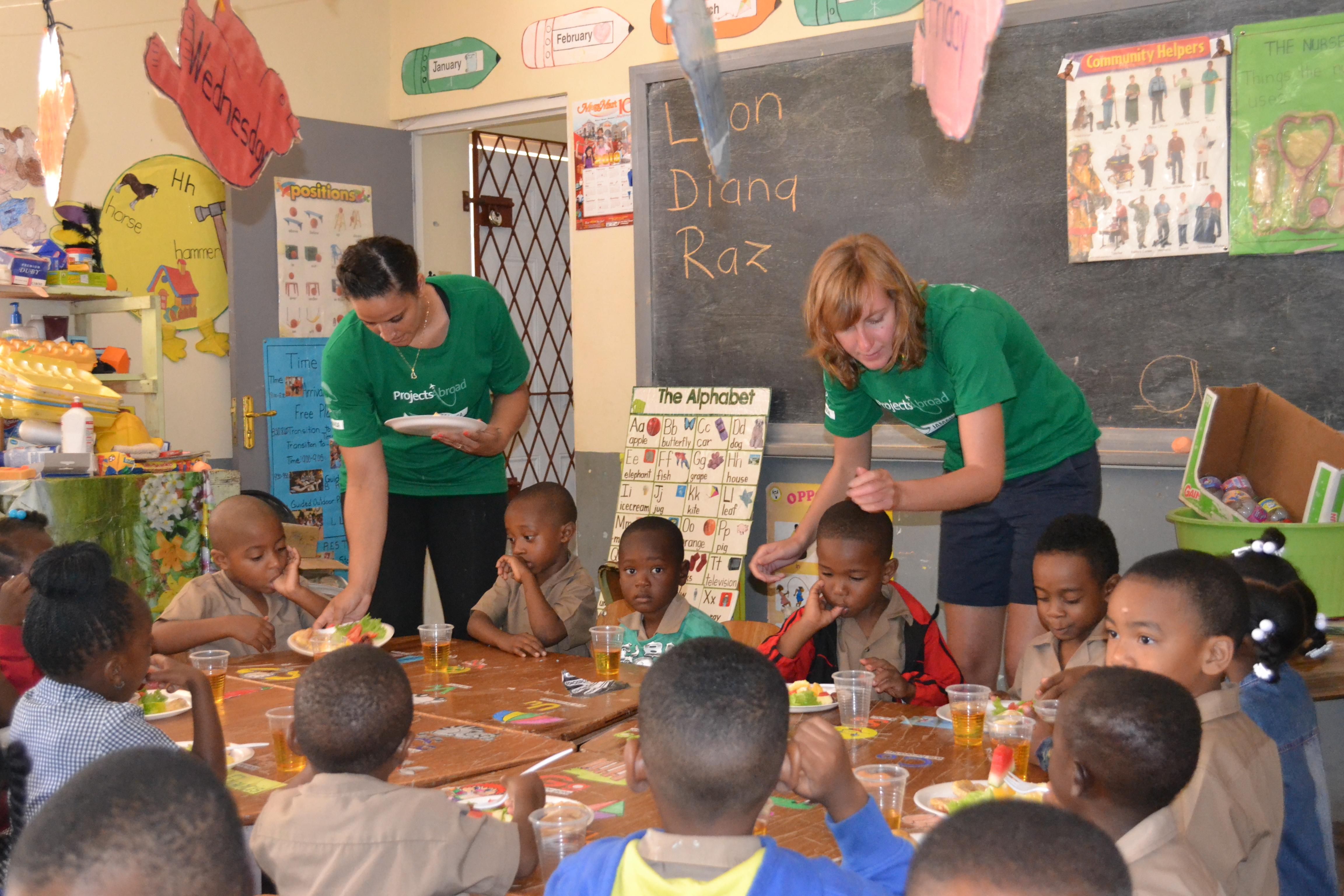 Projects Abroad female volunteers serve breakfast to kindergarten children during their Childcare Project in Jamaica. Two volunteers working with children in Jamaica serve breakfast to children at a kindergarten during a nutritional outreach.